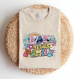 Puede incluir: Una camiseta beige con la frase "Ohana Means Family" en una fuente retro colorida. El dise&ntilde;o incluye un arco&iacute;ris, flores y estrellas. La camiseta est&aacute; doblada sobre una estera redonda tejida.