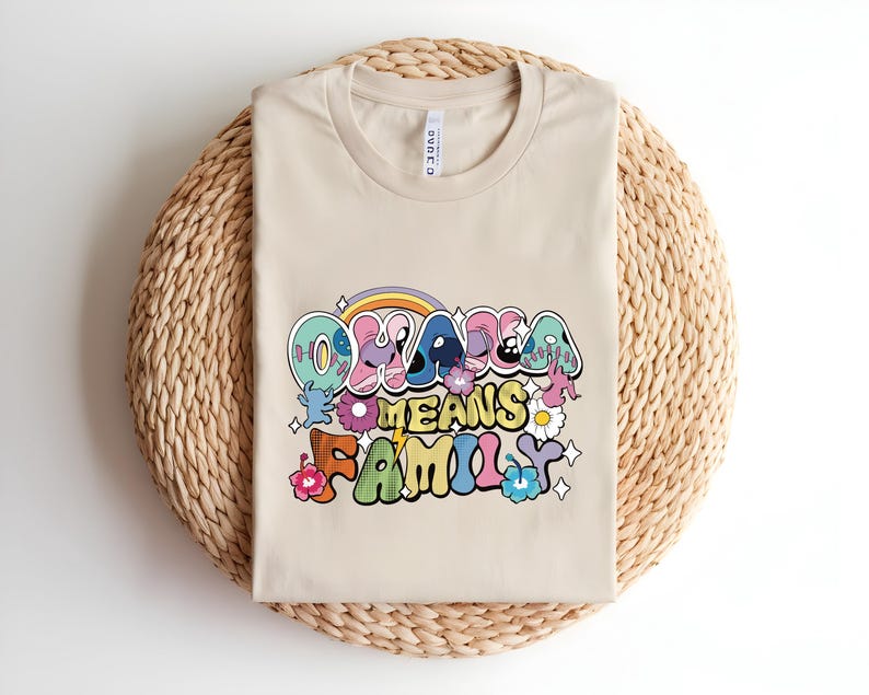 Puede incluir: Una camiseta beige con la frase "Ohana Means Family" en una fuente retro colorida. El dise&ntilde;o incluye un arco&iacute;ris, flores y estrellas. La camiseta est&aacute; doblada sobre una estera redonda tejida.