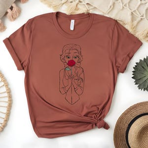 May include: A rust-colored t-shirt featuring a line drawing of a woman holding a red rose to her face. The design is centered on the front of the shirt. The shirt has short sleeves and a crew neck.