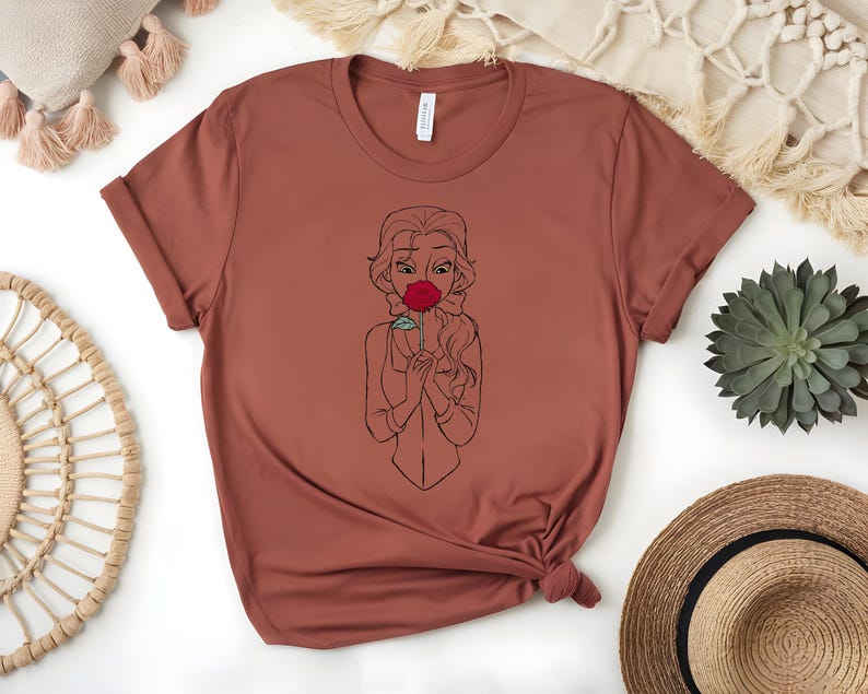 May include: A rust-colored t-shirt featuring a line drawing of a woman holding a red rose to her face. The design is centered on the front of the shirt. The shirt has short sleeves and a crew neck.