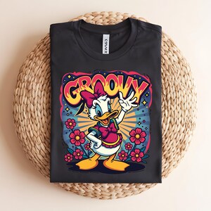 May include: Black t-shirt featuring a cartoon graphic of Daisy Duck with the word "GROOVY" in a colorful, retro font. Daisy is wearing a pink bow and vest, surrounded by flowers and a burst of rays.