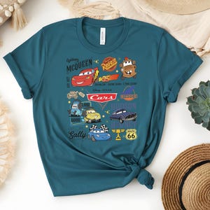 May include: Teal t-shirt featuring a colorful graphic of characters from the Disney Pixar Cars movie. The design includes Lightning McQueen, Mater, and other characters, along with the Cars logo and Route 66 signage. The shirt is made of soft material.
