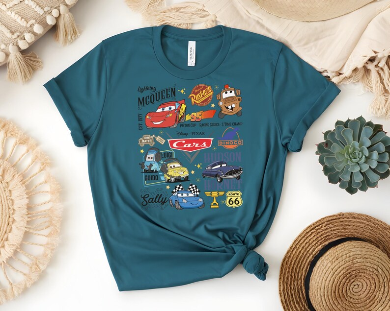 May include: Teal t-shirt featuring a colorful graphic of characters from the Disney Pixar Cars movie. The design includes Lightning McQueen, Mater, and other characters, along with the Cars logo and Route 66 signage. The shirt is made of soft material.