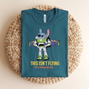 May include: Teal t-shirt featuring a cartoon character fusion of Stitch and Buzz Lightyear. The graphic shows Stitch in Buzz's spacesuit with the text "THIS ISN'T FLYING" and "This is falling with style."