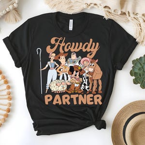 May include: Black t-shirt featuring a graphic of cartoon characters with the text "Howdy Partner" in a brown, western-style font. The design includes characters from a popular animated movie, with a cowboy theme.