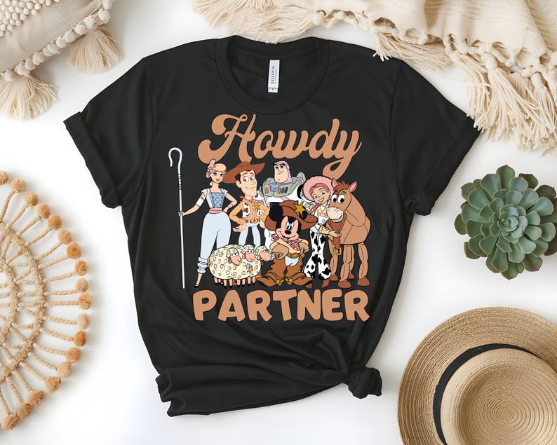 May include: Black t-shirt featuring a graphic of cartoon characters with the text "Howdy Partner" in a brown, western-style font. The design includes characters from a popular animated movie, with a cowboy theme.