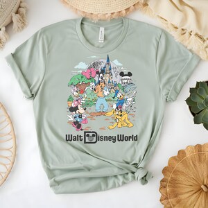 Puede incluir: Camiseta verde salvia con un gr&aacute;fico colorido de personajes y lugares emblem&aacute;ticos de Disney, incluyendo Mickey Mouse, Minnie Mouse y el castillo de Cenicienta. El texto "Walt Disney World" est&aacute; impreso en negro debajo.