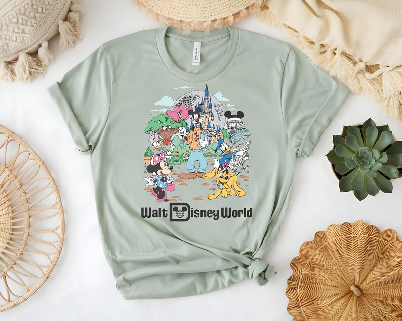 Puede incluir: Camiseta verde salvia con un gr&aacute;fico colorido de personajes y lugares emblem&aacute;ticos de Disney, incluyendo Mickey Mouse, Minnie Mouse y el castillo de Cenicienta. El texto "Walt Disney World" est&aacute; impreso en negro debajo.