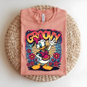 May include: Peach-colored t-shirt featuring a cartoon Daisy Duck design with the word "GROOVY" in a retro font. Daisy is wearing a pink bow and dress, surrounded by flowers and a burst of colorful rays.