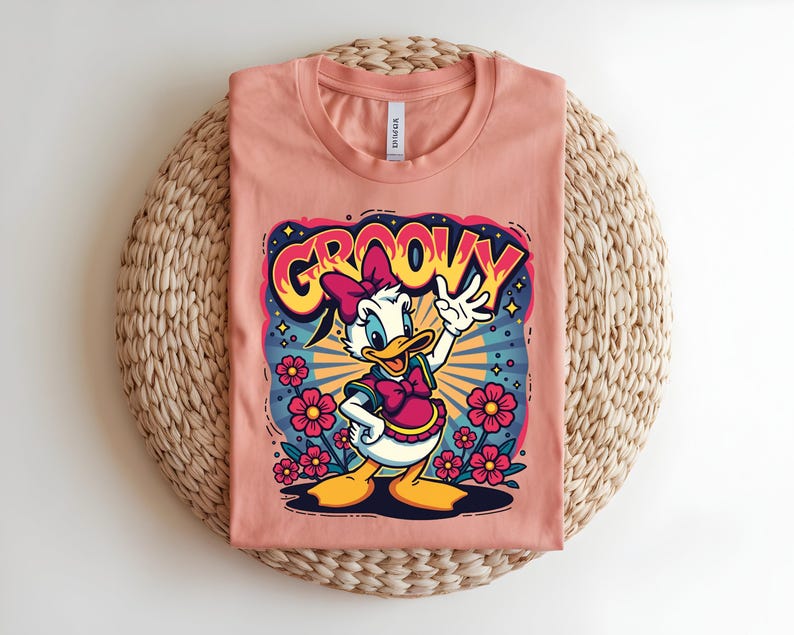 May include: Peach-colored t-shirt featuring a cartoon Daisy Duck design with the word "GROOVY" in a retro font. Daisy is wearing a pink bow and dress, surrounded by flowers and a burst of colorful rays.