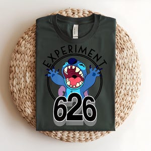 Pu&ograve; includere: T-shirt verde scuro con un personaggio dei cartoni animati blu, grandi orecchie e bocca aperta. Il testo "EXPERIMENT" &egrave; sopra il personaggio, con il numero "626" sotto. La maglietta &egrave; piegata su una superficie tessuta.