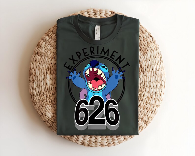 Pu&ograve; includere: T-shirt verde scuro con un personaggio dei cartoni animati blu, grandi orecchie e bocca aperta. Il testo "EXPERIMENT" &egrave; sopra il personaggio, con il numero "626" sotto. La maglietta &egrave; piegata su una superficie tessuta.