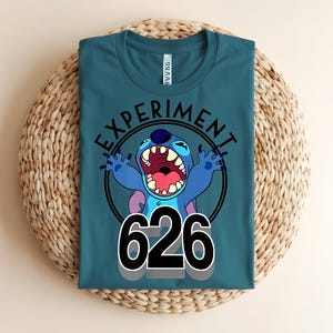 Pu&ograve; includere: T-shirt color teal con un personaggio dei cartoni animati e la scritta "EXPERIMENT 626". Il personaggio &egrave; blu con grandi orecchie e bocca aperta. Il testo &egrave; nero e il disegno &egrave; racchiuso in un cerchio nero.