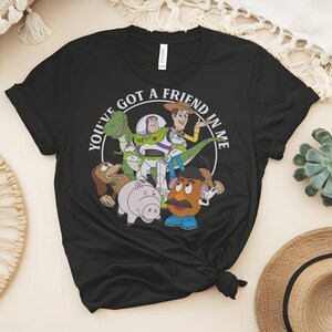 May include: Black t-shirt with a circular graphic featuring the text "YOU'VE GOT A FRIEND IN ME" and cartoon characters. The characters include a green dinosaur, Buzz Lightyear, Woody, and Mr. Potato Head. The shirt is made of a soft material.