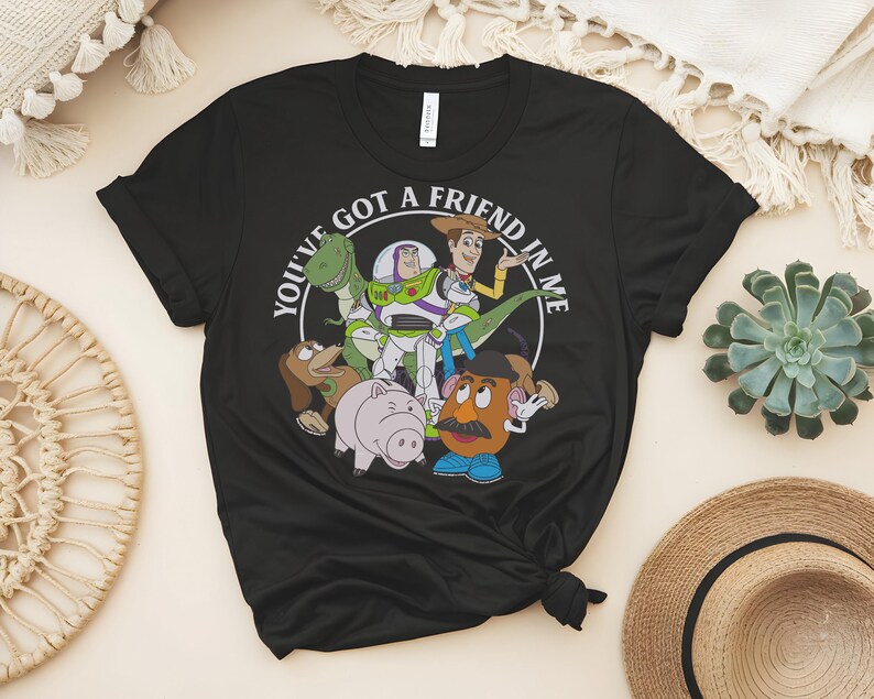May include: Black t-shirt with a circular graphic featuring the text "YOU'VE GOT A FRIEND IN ME" and cartoon characters. The characters include a green dinosaur, Buzz Lightyear, Woody, and Mr. Potato Head. The shirt is made of a soft material.