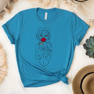 May include: A turquoise t-shirt featuring a line drawing of a woman holding a red rose to her face. The woman's hair is long and flowing, and she is wearing a simple outfit. The shirt is made of a soft material.