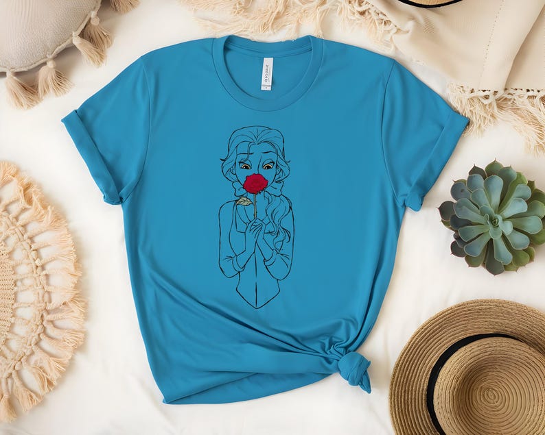 May include: A turquoise t-shirt featuring a line drawing of a woman holding a red rose to her face. The woman's hair is long and flowing, and she is wearing a simple outfit. The shirt is made of a soft material.