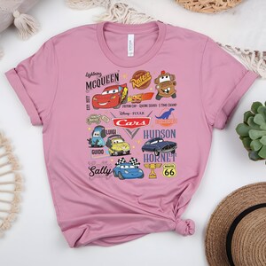 May include: A pink t-shirt featuring various cartoon car characters from the movie Cars. The design includes Lightning McQueen, Mater, and other characters, along with the text "Cars" and "Route 66".