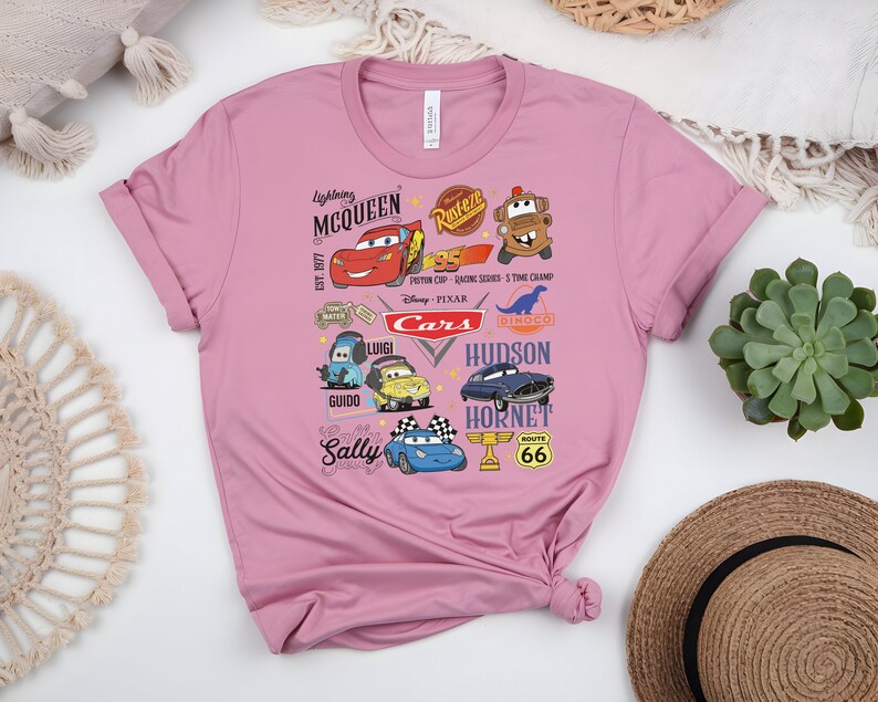 May include: A pink t-shirt featuring various cartoon car characters from the movie Cars. The design includes Lightning McQueen, Mater, and other characters, along with the text "Cars" and "Route 66".