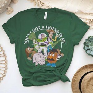 May include: A dark green t-shirt with a circular graphic that reads "YOU'VE GOT A FRIEND IN ME." The graphic features cartoon characters such as a dinosaur, Buzz Lightyear, Woody, and Mr. Potato Head. The t-shirt is knotted at the bottom.