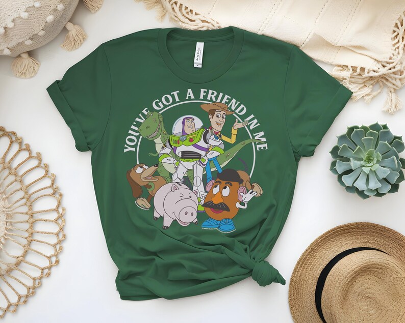 May include: A dark green t-shirt with a circular graphic that reads "YOU'VE GOT A FRIEND IN ME." The graphic features cartoon characters such as a dinosaur, Buzz Lightyear, Woody, and Mr. Potato Head. The t-shirt is knotted at the bottom.