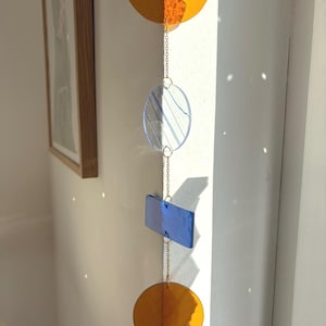 Puede incluir: Móvil colgante con formas geométricas en tonos naranja y azul. El móvil presenta una semicircunferencia, un círculo con líneas azules, un rectángulo y un círculo completo, todos conectados por delicadas cadenas doradas. Las formas son translúcidas.