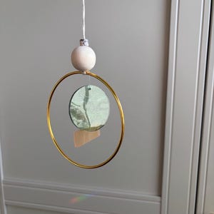 May include: A decorative hanging mobile with a gold-coloured metal ring, a light green circular element, and a small wooden shape. A wooden sphere and a white string are at the top. The mobile is suspended against a light grey background.
