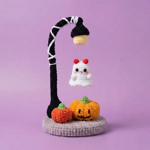 Puede incluir: Decoración de Halloween de ganchillo. Presenta una farola negra con detalles blancos, una calabaza naranja sonriente, una calabaza naranja más pequeña y un fantasma blanco con cuernos rojos. La base es un círculo gris.