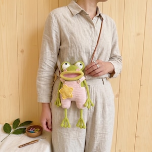 May include: A crochet frog-shaped crossbody bag featuring a green frog head with large white eyes, a pink mouth, and a light pink overall-style body with a yellow star appliqué on the front. The bag has long green arms and legs and a thin brown strap.