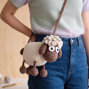 Puede incluir: Un bolso bandolera de ganchillo con forma de oveja, cuerpo color crema, cabeza y extremidades marrón oscuro y parte superior texturizada. El bolso presenta dos grandes ojos blancos con pupilas negras y una correa larga y fina de ganchillo marrón.