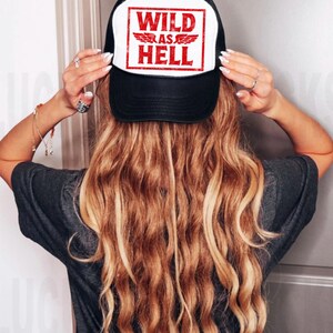 Op de afbeelding: Zwarte en witte truckerpet met de rode tekst "WILD AS HELL". De pet heeft een mesh achterkant en een gebogen klep. De persoon heeft lang, golvend blond haar.