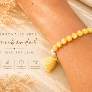 Könnte beinhalten: Ein gelbes Perlenarmband mit der Aufschrift "SUNKISSES" in weißen Buchstaben, getragen am Arm. Das Armband hat eine gelbe Quaste. Der Text im Bild lautet "PERSONALISIERTE Armbänder MIT HERZ. FÜR DICH."
