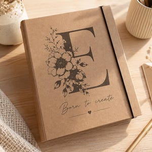 Op de afbeelding: Een rechthoekig notitieboek met een beige kraftpapieren kaft voorzien van een grote zwarte letter E versierd met bloemillustraties. Onder de letter staat de tekst Born to create in een cursief lettertype, gevolgd door een klein hartje.
