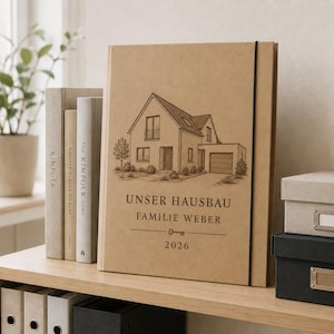 Op de afbeelding: Een beige hardcover plakboek met een zwarte elastische sluiting, voorzien van een donkerbruine lijntekening van een huis, de tekst "UNSER HAUSBAU FAMILIE WEBER 2026" en een klein sleutelpictogram op de voorzijde van de kaft.