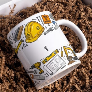 Puede incluir: Una taza de cerámica blanca con un patrón colorido de temática de construcción. El diseño incluye un casco amarillo con el texto Site Safety, una señal de Men At Work, una excavadora amarilla, un taladro, un nivel, planos y varias herramientas.
