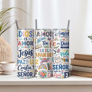 May include: Three cylindrical metal tumblers with lids and straws, featuring a white background covered in colorful religious text and illustrations including flowers, hearts, rainbows, butterflies, a cross, and an open book with Spanish phrases.