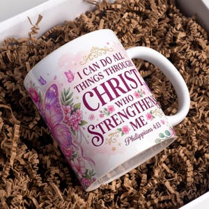 May include: A white ceramic mug featuring a purple butterfly, pink floral accents, and the text I CAN DO ALL THINGS THROUGH CHRIST WHO STRENGTHENS ME Philippians 4:13 printed in dark purple and gold lettering on the front surface.