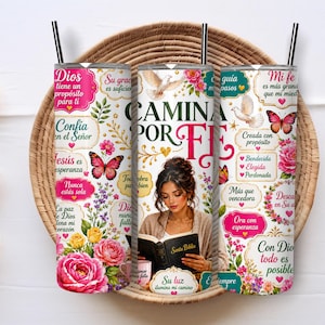 May include: A cylindrical tumbler with a straw featuring a white background, pink floral patterns, butterflies, and a central illustration of a person reading a book titled Santa Biblia. The text includes phrases like Camina por fe and Dios.