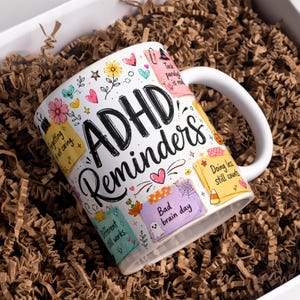以下が含まれることがあります： ADHD Remindersという黒い文字が書かれた白いセラミック製のマグカップ。付箋、ハート、花、幽霊のカラフルなイラストが描かれています。付箋にはForgetting is not caring、Doing less still counts、Bad brain dayといったテキストが含まれています。
