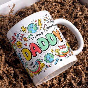 Peut inclure: Un mug en céramique blanche orné d'un motif graphique coloré avec le mot DADDY en grandes lettres. L'impression comprend des illustrations de fusée, de terre, d'éclairs, d'arcs-en-ciel et d'emojis, avec le texte so strong, super funny et cool dude.