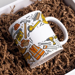 Puede incluir: Una taza de cerámica blanca con un patrón repetitivo y colorido de herramientas de construcción y equipo de seguridad, incluyendo un martillo, cinta métrica, taladro, sierra, llave inglesa, chaleco naranja, ladrillos y letreros con CAUTION MEN WORK.
