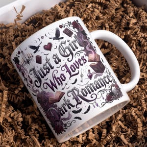 May include: A white ceramic mug featuring a dark-themed graphic with the text Just a Girl Who Loves Dark Romance. The design includes illustrations of ravens, open books, a chandelier, a heart, a potion bottle, and floral vines in shades of purple.