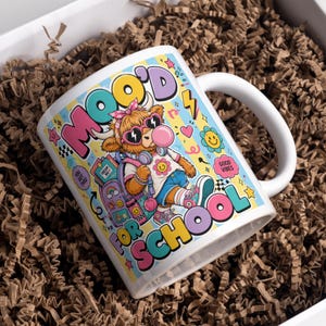 Peut inclure: Un mug en céramique blanche orné d'un graphisme coloré représentant une vache highland avec des lunettes de soleil, un bandeau et un sac à dos. Le texte indique MOO'D FOR SCHOOL, DREAM BIG, YOU GOT THIS et GOOD VIBES, avec des étoiles et fleurs.