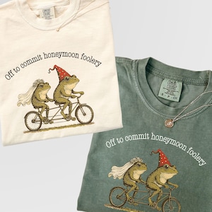 May include: A cream-colored t-shirt and a sage green t-shirt, both featuring a graphic of two frogs on a tandem bicycle. The frog in front wears a red party hat with white dots, and the frog in back wears a white veil. The text reads, Off to commit honeymoon foolery.