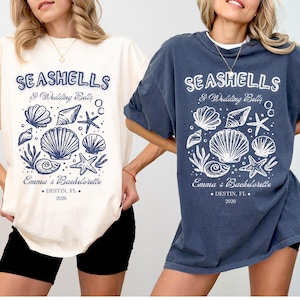 以下が含まれることがあります： クリーム色とネイビーブルーの2枚のTシャツ。貝殻とヒトデのグラフィックが特徴です。「SEASHELLS & Wedding Bells」と「Emma's Bachelorette, Destin, FL, 2026」の文字。カジュアルでオーバーサイズのシャツです。