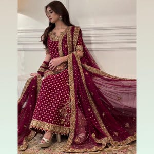 May include: A maroon embroidered salwar kameez ensemble with gold embellishments. The outfit includes a long tunic, matching pants, and a sheer dupatta. The intricate gold embroidery trims the neckline, cuffs, and hem. The outfit is worn with gold sandals.