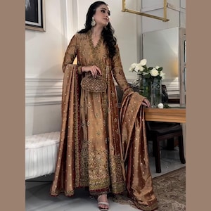 Könnte beinhalten: Ein langes, fließendes, goldfarbenes Abendkleid mit aufwendigen Stickereien und Verzierungen. Das Kleid hat lange Ärmel und einen passenden Schal. Eine kleine, perlenbesetzte Handtasche und silberne Sandalen vervollständigen das Ensemble. Geeignet für besondere Anlässe.