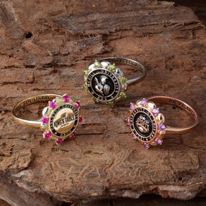 May include: Three metal rings featuring circular faces with engraved text and colorful stones. One ring has pink stones and reads Carleton University The King's Table, another has green stones and McGill University, and the third has purple stones and University of Chicago.