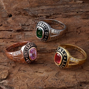 May include: Three class rings in silver, rose gold, and yellow gold tones. The silver ring features a green stone and text reading UNIVERSITY OF CHICAGO. The rose gold ring has a pink stone and UNIVERSITY text. The yellow gold ring has a red stone and YALE UNIVERSITY text.