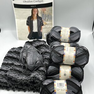 May include: A collection of gray, black, and white variegated yarn skeins with a paper pattern sheet titled Obsidian Cardigan. The yarn features a gradient color scheme, and the pattern sheet displays a photograph of a finished knit cardigan.